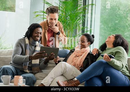 Verschiedene Freunde treffen sich auf dem Sofa im Wohnzimmer und sehen ein Tablet mit Tassen auf dem Couchtisch Stockfoto
