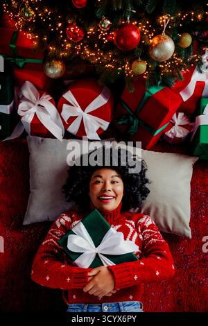 Fröhliche Frau, die ein festliches Geschenk umarmt, umgeben von farbenfrohen Weihnachtsdekorationen und hell verzierten Geschenken Stockfoto