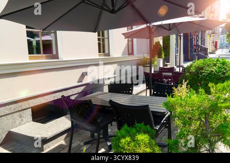 Sommerblick auf ein leeres Straßencafé oder eine Restaurantterrasse mit dunklen Tischen und Stühlen, die von großen schwarzen Sonnenschirmen im hellen Sonnenlicht beschattet werden Stockfoto