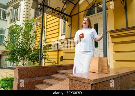 Selbstbewusste junge Frau in Übergröße, die mit Kaffee zum Mitnehmen und Papiertüten in der Nähe des Eingangs eines hellgelben Gebäudes steht Stockfoto