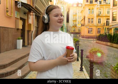 Eine schöne Frau in Übergröße macht einen Spaziergang in der sonnigen europäischen Stadt und hört ihre Lieblingsmusik bei einer Tasse Kaffee zum Mitnehmen Stockfoto
