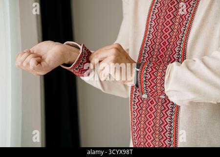 Eine Nahaufnahme zeigt einen Mann, der die Manschette eines beigefarbenen Leinenhemdes sorgfältig anpasst, das reich mit roten und schwarzen ukrainischen Vyshyvanka-Mustern bestickt ist. Stockfoto