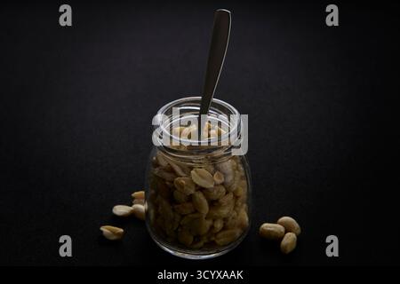 Vertikales Foto von Erdnüssen im Glasgefäß mit Löffel auf schwarzer Oberfläche. Minimalistisches Dark Food-Konzept, das gesunden veganen Protein Snack betont. Stockfoto
