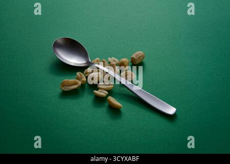 Ein silberner Löffel gefüllt mit Erdnüssen auf einem lebhaften grünen Hintergrund unter weichem Licht. Die farbenfrohe, minimalistische Einrichtung betont sauberes Essen und Natu Stockfoto