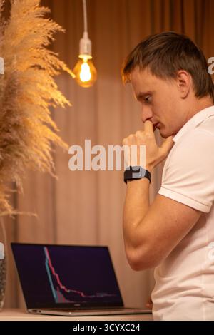 Nachdenklicher junger Mann im Spannungsfeld Analyse von Marktdiagrammen und Handelsdaten für Kryptowährungen auf Laptop im modernen Home Office Workspace. Unternehmen, Mann. Stockfoto
