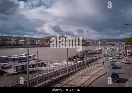 Blick auf die Donau in Budapest, mit Booten, die am Pier angedockt sind, Autos, die am Ufer vorbeifahren, und das Budaer Schloss auf dem Hügel. Stockfoto