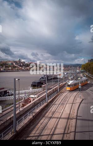 Blick auf die Donau in Budapest, mit Booten, die am Pier angedockt sind, Autos, die am Ufer vorbeifahren, und das Budaer Schloss auf dem Hügel. Stockfoto