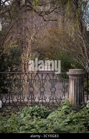 Ein Gartenweg umgeben von üppigem Grün und Bäumen, mit einem kunstvollen schmiedeeisernen Zaun und einer Steinsäule im Vordergrund. Stockfoto