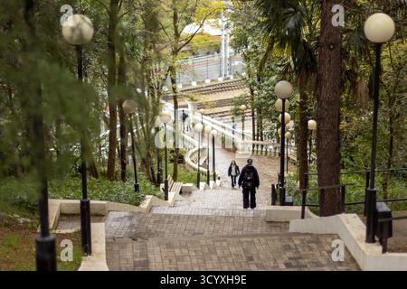 Kopfsteinpflastertreppen im üppigen Park. Kopfsteingepflasterte Treppen, die sich durch einen üppigen Stadtpark mit hohen Bäumen, Vintage-Lampen und entfernten Bahngleisen hinunter führen Stockfoto