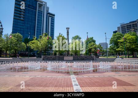 Wasserstrahlen erheben sich aus dem Springbrunnen der Olympischen Ringe im Centennial Olympic Park, eingerahmt von modernen Hochhäusern, Bäumen und amerikanischen Flaggen unter klarem Himmel Stockfoto