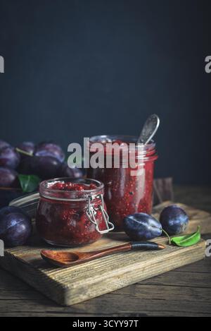 Pflaumenmarmelade in Glasgläsern mit Reifen Pflaumen auf dunklem Hintergrund Stockfoto