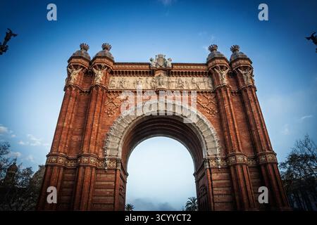 Tiefwinkelansicht des Arc de Triomf in Barcelona, Spanien. Das rote Ziegelsteindenkmal mit Friesen und einem großen Bogen steht vor einem klaren blauen Himmel. Stockfoto
