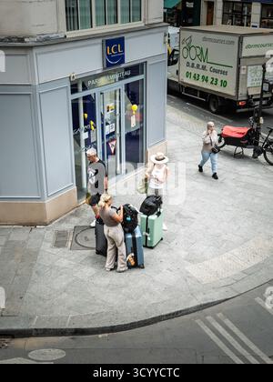 PARIS, FRANKREICH - 26. JUNI 2025: Touristen mit Koffern halten an einer Ecke in Le Marais, Paris, und überprüfen die Wegbeschreibung in der Nähe von Geschäften und Verkehr. erfassen Stockfoto
