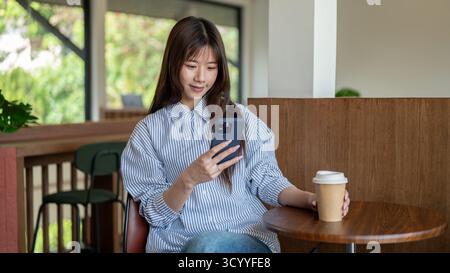 Lächelnde asiatische Frau in blauem Hemd, die auf ihr Handy schaut und sich eine Kaffeetasse schnappt, während sie an einem hölzernen Hocker im Café sitzt. Kühlung Im Freien. Stockfoto