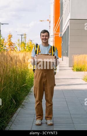 Ein freundlicher Kuriermann lächelnd, präsentiert einen Paketkasten, steht auf einem Bürgersteig, liefert Pakete an Kunden in einem städtischen Umfeld, bietet Expressversand und Logistikservice Stockfoto
