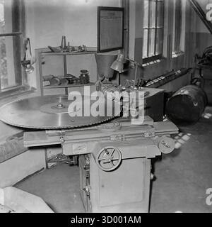 Dieses Archivfoto von Sloviansk aus dem Jahr 1966 zeigt eine Spezialmaschine in einer Werkstatt in der Armature and Insulator Factory (AIF). Das Bild zeigt eine Schleifmaschine für Kreissägeblätter, eine wichtige Stützausrüstung. Ein sehr großes, gezahntes Sägeblatt ist horizontal montiert und wird von einer kleineren Schleifscheibe gewartet, die von einem Arbeitsscheinwerfer positioniert wird. Der Innenraum der Werkstatt mit Regalen und Fenstern ist sichtbar. Diese Aufnahme verdeutlicht die wichtige Wartung und technische Qualifikation, die zur Unterstützung der Schwerindustrie erforderlich ist, ein Dokument des friedlichen Donbass Stockfoto