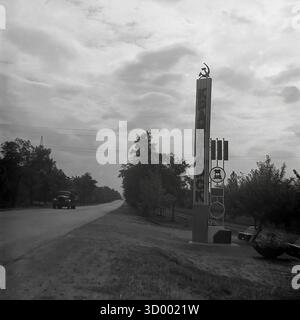 Dieses Archivfoto aus dem Jahr 1966 zeigt das Haupteingangsschild (Stele) nach Kramatorsk, Ukrainische SSR, das die Stadtgrenze mit Sloviansk markiert. Der hohe, modernistische Pylon trägt den Namen der Stadt „КРАМАТОРСК“ (KRAMATORSK) in vertikaler kyrillischer Schrift, gekrönt mit dem sowjetischen Wappen (Hammer und Sichel). Ein nebenan angebrachtes Schild zeigt Industriesymbole (ein Zahnrad), die die Schwerindustrie der Stadt widerspiegeln. Ein Oldtimer fährt auf der Autobahn. Dieses Wahrzeichen repräsentiert den Stolz eines großen Industriezentrums, ein Schlüsseldokument des friedlichen Donbass Stockfoto