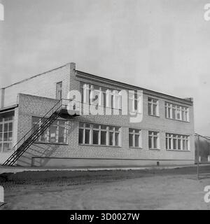 Dieses Archivfoto aus dem Jahr 1966 von Sloviansk, der ukrainischen SSR, zeigt ein typisches zweistöckiges öffentliches Gebäude aus der Sowjetzeit. Das Gebäude wurde aus weißem Silikatstein im funktionalen Stil der 1960er Jahre gebaut und verfügt über große, sich wiederholende Fenster. Seitlich ist eine Außentreppe aus Metall für den Notausstieg angebracht. Das unbefestigte Gelände um das Gebäude herum weist Reifenspuren auf, was möglicherweise auf eine kürzliche Baumaßnahme hindeutet. Dieses Bild fängt die gemeinsame städtische Architektur jener Zeit ein, ein Dokument der alltäglichen Umgebung des friedlichen Donbas Stockfoto