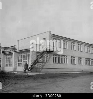 Dieses Archivfoto aus dem Jahr 1966 von Sloviansk, der ukrainischen SSR, zeigt ein typisches zweistöckiges öffentliches Gebäude aus der Sowjetzeit. Das Gebäude wurde aus weißem Silikatstein im funktionalen Stil der 1960er Jahre gebaut und verfügt über große, sich wiederholende Fenster. Seitlich ist eine Außentreppe aus Metall für den Notausstieg angebracht. Das unbefestigte Gelände um das Gebäude herum weist Reifenspuren auf, was möglicherweise auf eine kürzliche Baumaßnahme hindeutet. Dieses Bild fängt die gemeinsame städtische Architektur jener Zeit ein, ein Dokument der alltäglichen Umgebung des friedlichen Donbas Stockfoto
