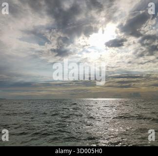 Der Himmel ist bewölkt und die Sonne scheint durch die Wolken. Das Meer ist ruhig und das Wasser reflektiert die Sonne Stockfoto