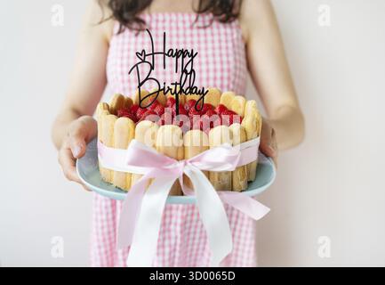Ein Mädchen in einem rosa Kleid hält einen Erdbeermousse-Kuchen in den Händen, mit frischen Erdbeeren und der Inschrift Happy Birthday Stockfoto