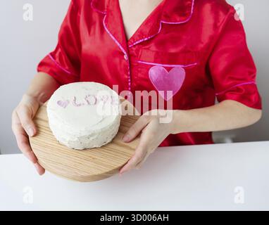 Ein Mädchen in einem roten Seidenhemd mit Herz hält einen weißen Kuchen mit der Aufschrift LIEBE auf einem Holzbrett. Vorbereitung auf den Valentinstag Stockfoto