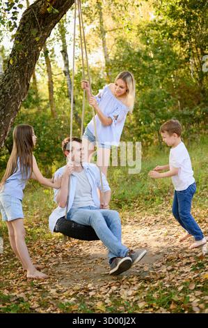Die Familie spielt gern draußen, mit Vater auf selbstgemachter Reifenschaukel, Mutter hinter ihm, Junge und Mädchen schwingt ihre Eltern. Freizeit fa Stockfoto