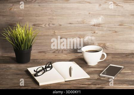 Arbeitstisch mit Notizblock, Kaffeetasse, Stift, Smartphone, Brille, Pflanztopf. Holztischhintergrund mit Kopierraum oben. Instant Photo Vintage spl Stockfoto