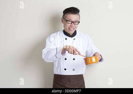 Porträt eines reiferen asiatischen Küchenchefs in einheitlicher Küche, stehend auf schlichtem Hintergrund mit Schatten, Kopierraum an der Seite Stockfoto