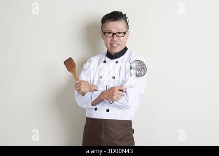 Porträt eines selbstbewussten, reiferen asiatischen Küchenchefs aus den 50er Jahren in einheitlichen Händen, der Küchenwerkzeuge hält, auf schlichtem Hintergrund mit Schatten steht, Kopierraum Stockfoto
