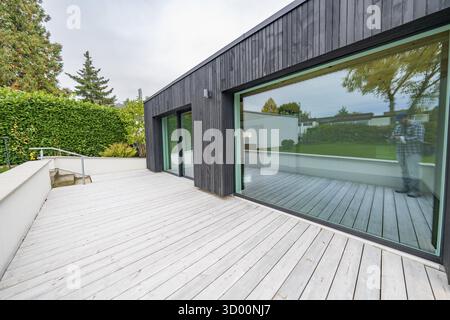 Minimalistischer Anbau mit schwarzer Holzfassade und großer Glasfront öffnet sich auf eine breite Holzterrasse im ruhigen Garten, Hausrenovierung, Klimaanlage Stockfoto