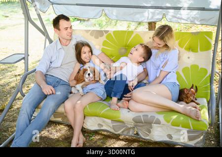 Familie und Kinder entspannen sich im Freien mit Hunden. Eltern und Kinder sitzen auf einer Gartenschaukel, schaukeln, plaudern und teilen fröhliche Momente im Hof. Konzept Stockfoto
