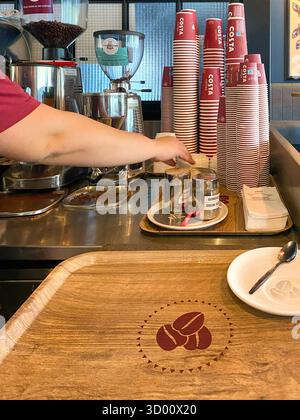 Costa Coffee Barista bereitet Getränke an einer Kaffeestation zu, mit gestapelten Tassen, Kaffeemühle und Holztablett mit Kaffeebohne Logo Stockfoto