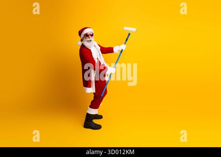 Charismatischer Weihnachtsmann, der die Weihnachtszeit feiert, während er mit einem blauen Malerroller auf gelbem Hintergrund posiert Stockfoto