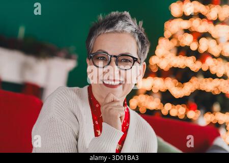 Lächelnde ältere Frau in festlicher Weihnachtsstimmung drinnen mit leuchtenden Weihnachtsbaumlichtern und gemütlicher Dekoration Stockfoto