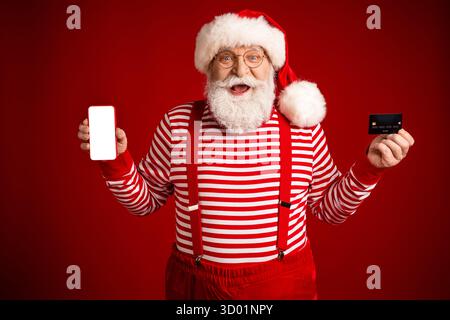 Fröhliche Weihnachtsmann-Figur in rot gestreiftem Hemd und Hosenträgern hält ein leeres Smartphone und eine Karte vor festlichem rotem Hintergrund Stockfoto