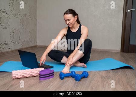 Lächelnde Frau in Sportbekleidung sitzt auf einer Yogamatte mit Laptop und Kurzhanteln, macht Heimtraining und Online-Fitness-Trainingskonzept Stockfoto