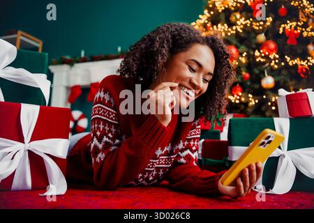 Glücklicher Feiertagsmoment, als eine junge Frau in einem roten Pullover auf einem hellgelben Telefon neben einem Baum voller Lichter und gewickelter Geschenke plaudert Stockfoto