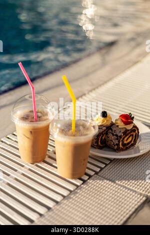 Zwei Eiskaffee-Drinks mit bunten Strohhalmen sitzen an einem hellen, sonnigen Nachmittag neben einem Teller mit Schokoladenrollen in der Nähe eines glitzernden Pools. Stockfoto