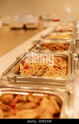 Eine Auswahl an Nudelgerichten mit verschiedenen Saucen und Zutaten ist auf einem langen Tisch in Metalltabletts untergebracht. Dieses Buffet lädt Gäste zum Servieren ein Stockfoto