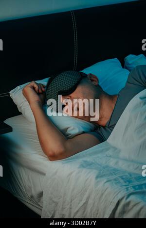 In einem Schlafzimmer liegt ein Mann auf seiner Seite mit einer Schlafmaske, die seine Augen bedeckt, auf einem Kissen und in ein weißes Tuch gewickelt, ein Arm über dem Pfol gebeugt Stockfoto
