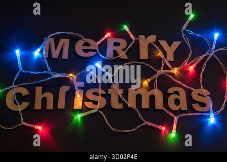 Weihnachtskarte mit schwarzem Hintergrund, ein kleiner Baum mit cremefarbenen Holzbuchstaben, die Frohe Weihnachten sagen, und bunte LED-Lichter. Stockfoto