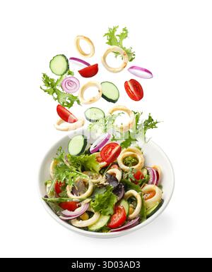 Tintenfischringe und andere Zutaten, die in eine Schüssel mit Salat vor weißem Hintergrund fallen Stockfoto