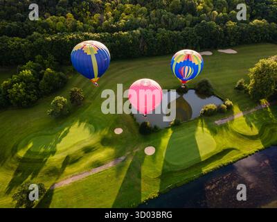 Top-down-Luftaufnahme von drei lebendigen Heißluftballons, die während der goldenen Stunde aus einem grünen Park steigen und bunte Muster einfangen Stockfoto