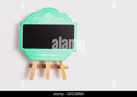 Eine dekorative, grün gerahmte Mini-Tafel mit kunstvollen weißen Details, die auf einer Staffelei aus Holz vor einem sauberen weißen Hintergrund steht. Stockfoto