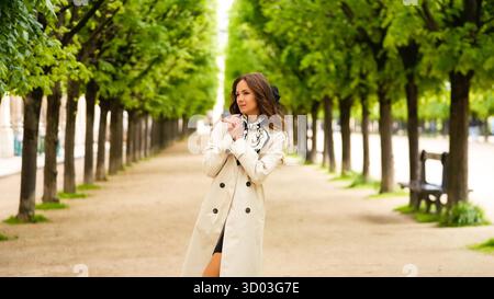 Elegante Frau im Trenchcoat und Schal, die an einem Frühlingstag unter einem Baumdach in der Nähe des Palais Royal in Paris spaziert. || Modell freigegeben Stockfoto