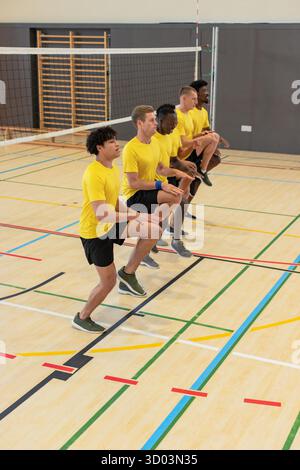 Verschiedene männliche Mannschaftskameraden führen synchronisierte Kniehegelungen in der Sporthalle mit Volleyballnetzpfosten durch. Athleten, Teamwork, Fitness, Training, Gymnasium, Übung, Kameradschaft || Model veröffentlicht Stockfoto