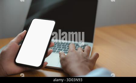 Nahaufnahme eines Handheld-Smartphones mit leerem weißen Bildschirm und Laptop auf Holzschreibtisch. Perfekt für Mockup, Design von mobilen Apps, digitales Geschäft und vieles mehr Stockfoto