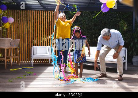 Großeltern und Enkelin feiern mit bunten Streamern auf der sonnigen Terrasse. Familie, Feier, Festlichkeit, Glück, Mehrgenerationen, Outdoor || Modell veröffentlicht Stockfoto