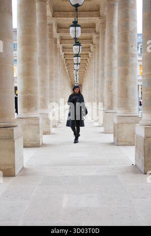 Eine Frau in schickem Winteroutfit spaziert durch die großen Säulen des Palais Royal in Paris und verkörpert Pariser Eleganz und Stil in einer historischen Umgebung. Hochwertiges Foto || Modell veröffentlicht Stockfoto
